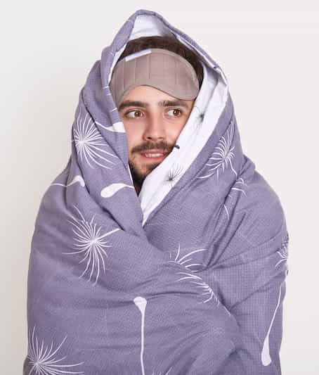 Bearded guy with sleeping mask on forehead looking aside, being wrapped in blanket, copy space advertisement or promotion text, unshaven man feels comfort.